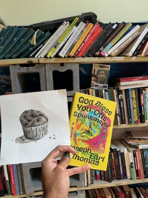 A hand holds a drawing of a muffin and a copy of "God Bless You, Otis Spunkmeyer," by Joseph Earl Thomas.