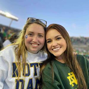 Two smiling students pose for a photo in the stands of Notre Dame Stadium. One student wears a gray sweatshirt with "NOTRE DAME" and the other wears a green Notre Dame Law School sweatshirt. A partially visible stadium light and other fans are in the background.