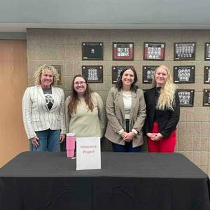 Four individuals stand behind a table draped in a black tablecloth. A sign on the table reads "Innocence Project." On the wall behind them are framed photos of graduating classes from the University of Notre Dame School of Law. A wooden door is visible to the left.