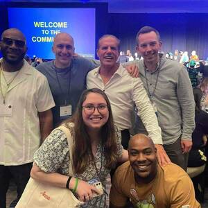 Six individuals pose for a photo at a conference. Four men stand shoulder to shoulder in the back, while a woman and another man kneel in the foreground. A large screen behind them displays a welcome message to the “Community Network”. Attendees are seated at tables in the background.