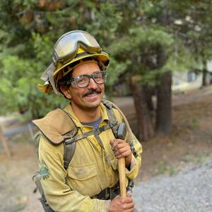 A firefighter in a yellow uniform and protective gear, including a helmet and goggles, smiles while holding an axe.