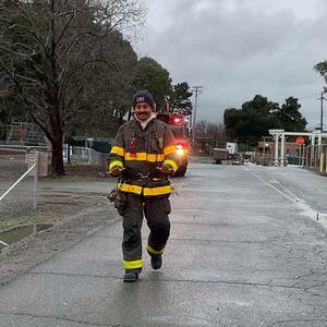Firefighter in dark uniform with yellow stripes and a "Fire Rescue" beanie walks toward the camera, gesturing with his hands. A fire truck with flashing lights is visible in the background.