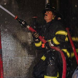 A firefighter in dark bunker gear with yellow reflective stripes kneels on wet steps, aiming a red firehose with an orange nozzle. Water sprays from the hose.