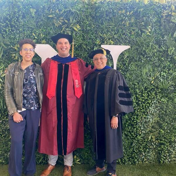Three smiling individuals, a woman in a dark jacket and two men in academic caps and gowns, pose before a green foliage wall with a white ND logo. One man wears a maroon gown with a blue hood; the other a black gown with a blue hood.