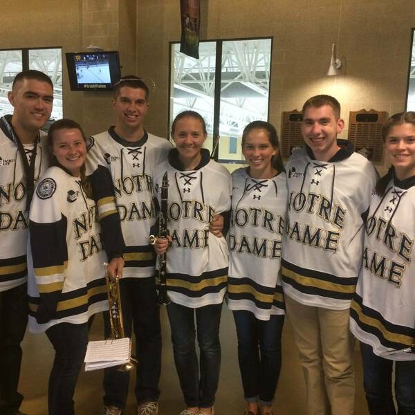 Seven smiling Notre Dame students, four women and three men, wear white, navy, and gold hockey jerseys in an indoor arena. Two hold a trumpet with sheet music and a clarinet. A TV screen in the background shows a hockey game.