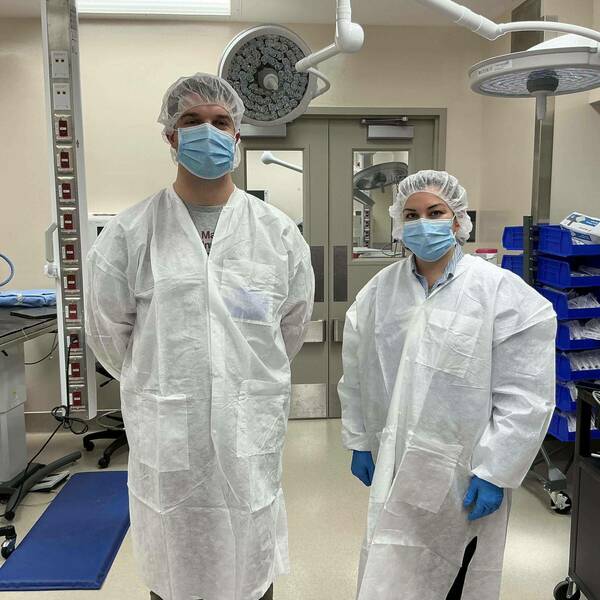 A man and woman in white lab coats, hairnets, and blue masks stand in a medical room. Surgical lights hang from the ceiling, and blue bins filled with supplies are on the right. The woman also wears blue gloves.