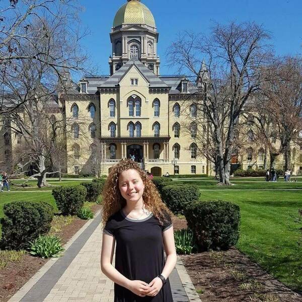 Young woman with curly hair smiles in a black dress on a sunny day in front of Notre Dame's Main Building with the Golden Dome.