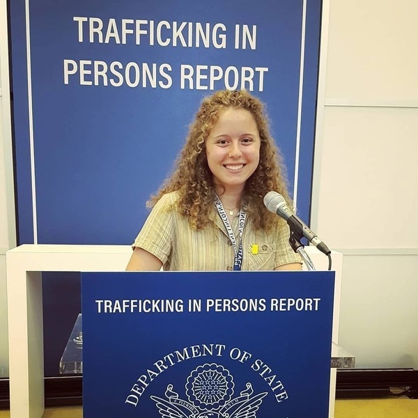 Smiling woman with curly hair at a podium with a microphone. Blue signs read "Trafficking in Persons Report."