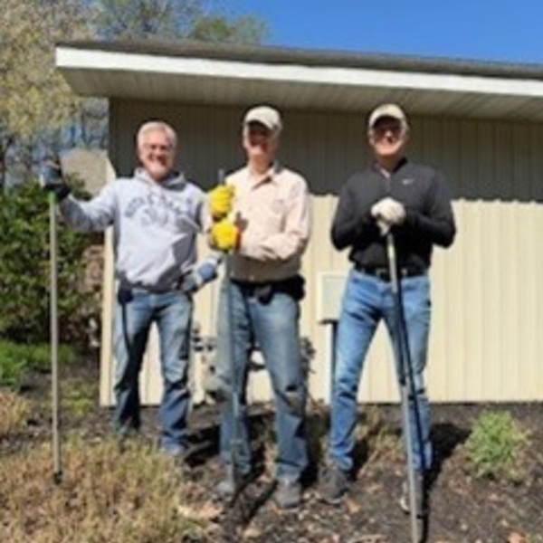 Three smiling men, one in a Notre Dame sweatshirt, hold gardening tools in a mulched bed on a sunny day.