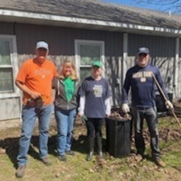 Four smiling adults stand in a yard with fallen leaves. Two wear Notre Dame apparel, one holds a rake near a leaf bag.
