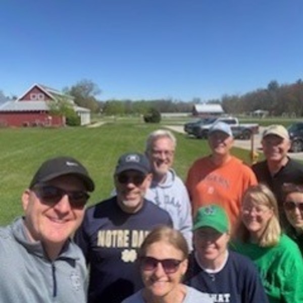 Nine smiling adults, some wearing Notre Dame gear, pose for a selfie outdoors in a grassy field with red barns.