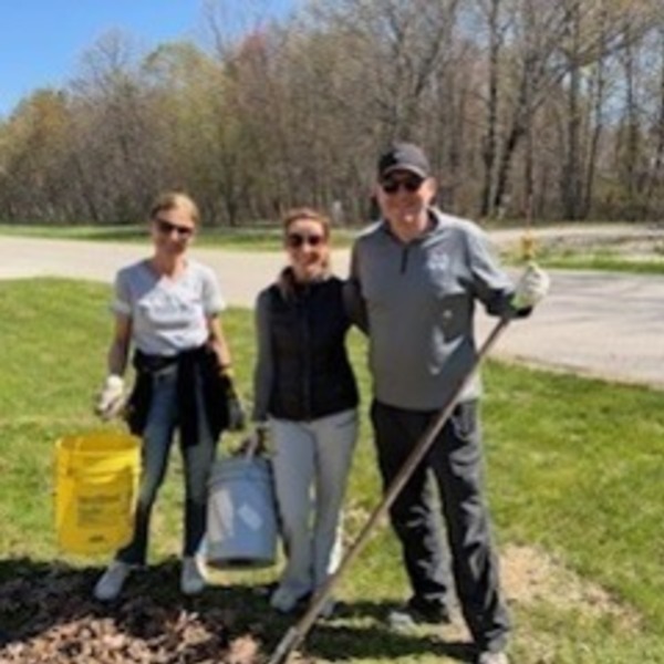 A man and two women, smiling, wearing sunglasses and work gloves, hold a rake and buckets while cleaning leaves outdoors.