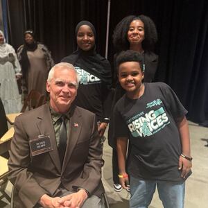 Smiling man in wheelchair, two smiling girls and a smiling boy wearing 'Rising Voices' shirts on a stage.